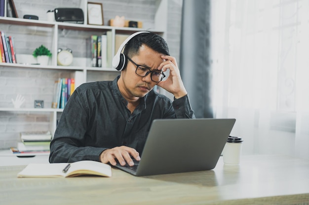 L'uomo freelance asiatico indossa occhiali e cuffie con depressione stressante momento triste mentre lavora in videoconferenza riunione sul laptop Uomo depresso triste serios lavorando a casa Lavoro ovunque concetto