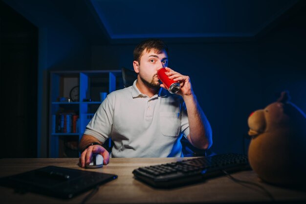 L'uomo freelance lavora di notte su un computer a casa e beve una cola guardando