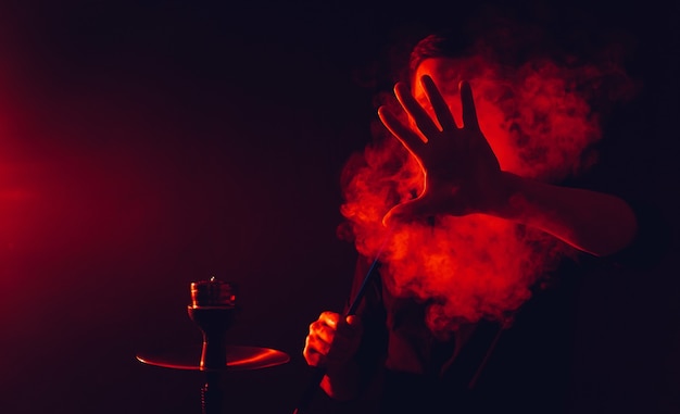 L'uomo fuma un narghilè in un bar e soffia una grande nuvola di fumo con luci al neon rosse