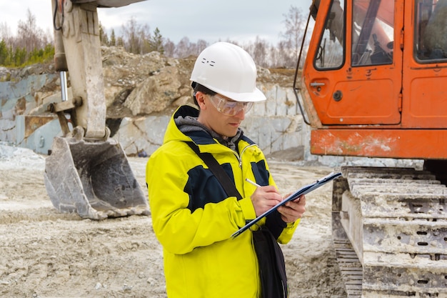 L'uomo geologo o un ingegnere minerario scrive qualcosa in una custodia per mappe in mezzo a una cava con attrezzature da costruzione