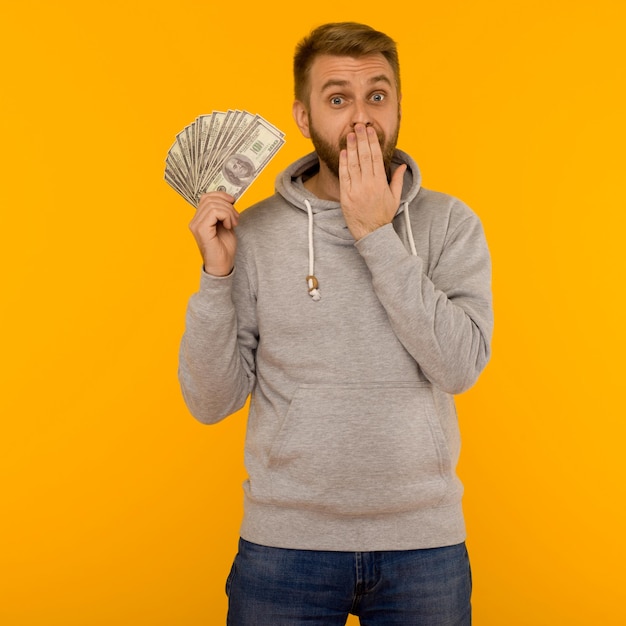 L'uomo gioioso con una felpa con cappuccio grigia tiene in mano dollari che coprono la bocca con la mano su un'immagine di sfondo gialla