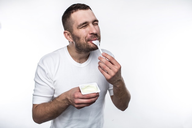 L'uomo giovane e forte sta mangiando lo yogurt. Lo sta facendo con piacere. Isolato su sfondo bianco