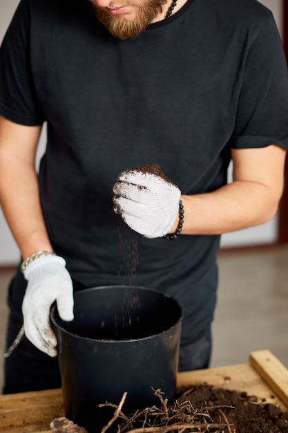 L'uomo ha messo il terreno in vaso nero sul tavolo di legno, trapianto di piante da appartamento, hobby e tempo libero, giardinaggio domestico.