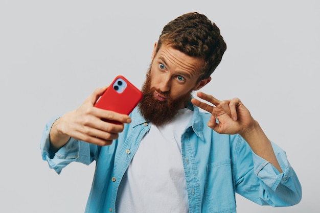 L'uomo hipster con il telefono in mano blogger sorride su sfondo grigio in camicia blu e maglietta bianca che parla al telefono e selfie