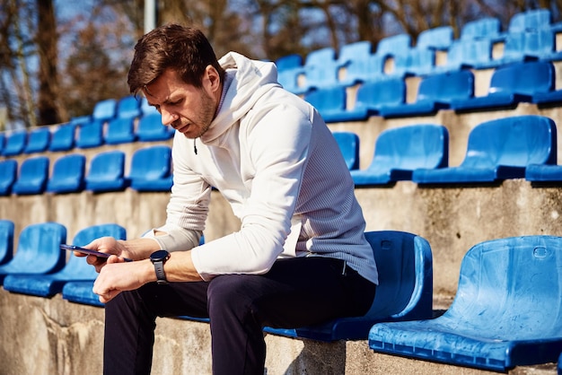 L'uomo in abbigliamento sportivo seduto sul sedile dello stadio e sfogliando l'orologio sportivo maschio dello smartphone controlla il fitness