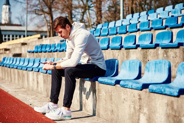 L'uomo in abbigliamento sportivo seduto sul sedile dello stadio e sfogliando l'orologio sportivo maschio dello smartphone controlla il fitness