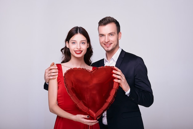 L'uomo in abito e la donna in abito rosso tengono un pallone a forma di cuore rosso su sfondo bianco