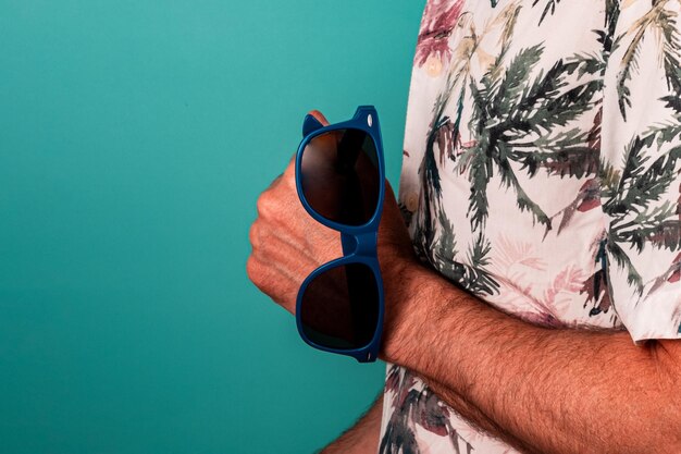 L'uomo in camicia hawaiana tiene in mano occhiali da sole blu con uno sfondo verde