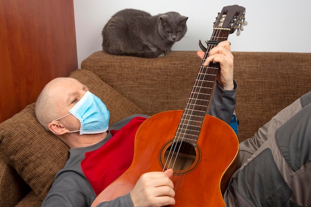 L'uomo in quarantena a casa con una maschera medica sul viso giace sul divano e suona la chitarra. Riposo durante l'epidemia di coronavirus.