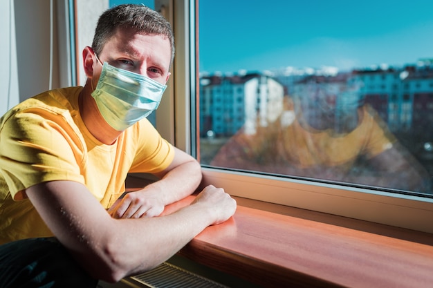 L'uomo in quarantena si siede a casa in una maschera e guarda fuori dalla finestra in una giornata di sole. Concetto di coronavirus.