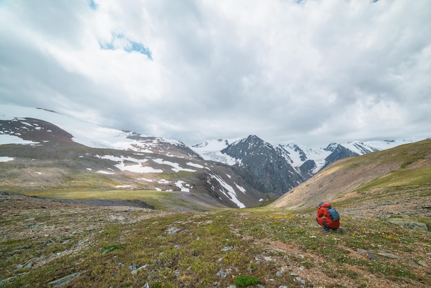 L'uomo in rosso con la fotocamera scatta la vista con un'alta catena montuosa innevata con cime affilate e ghiacciai sotto il cielo nuvoloso grigio Il turista fotografa il paesaggio con grandi montagne di neve con tempo variabile