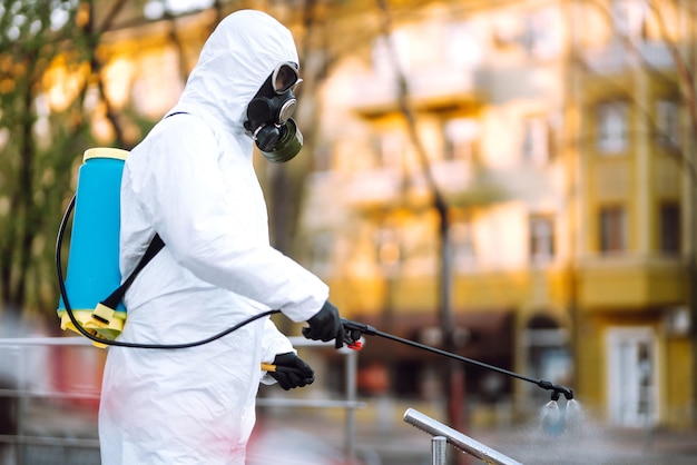 L'uomo in tuta protettiva e maschera spruzza disinfettante sulla ringhiera nel luogo pubblico Covid 19