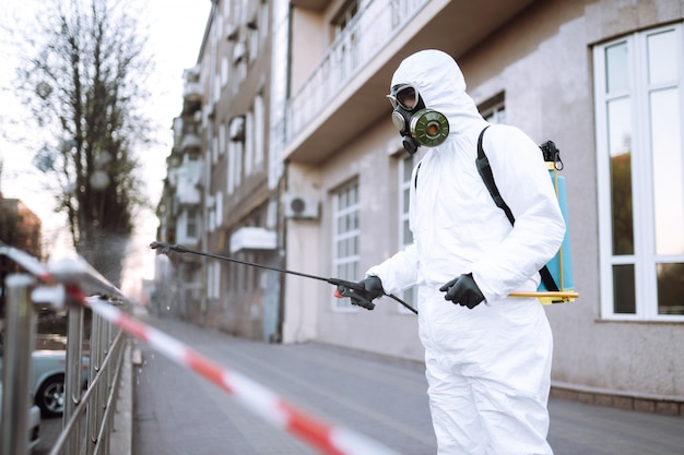 L'uomo in tuta protettiva e maschera spruzza il disinfettante sulla ringhiera nel luogo pubblico vuoto all'alba nella città di quarantena. Covid 19. Concetto di pulizia.