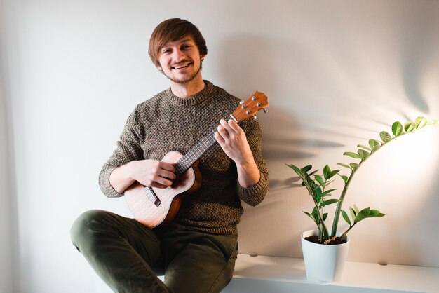 L'uomo in un maglione marrone sorride e gioca sulle ukulele mentre è seduto a casa.