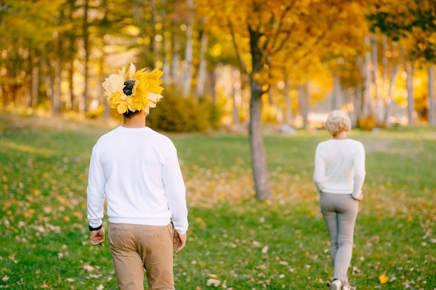 L'uomo in una corona di foglie gialle segue la donna in abiti leggeri nella vista posteriore della foresta di autunno