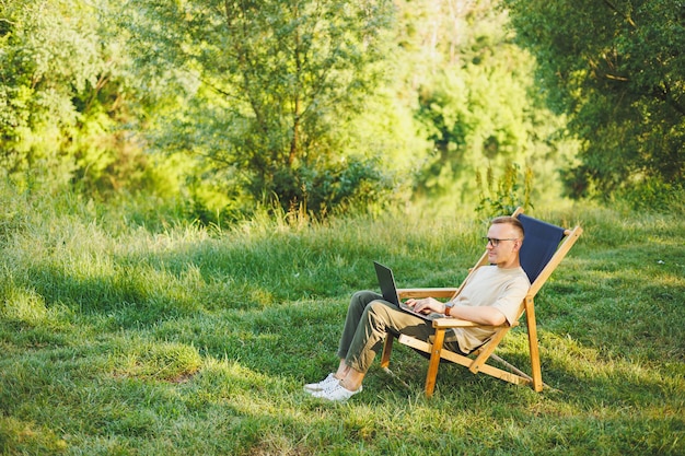 L'uomo libero professionista si siede su una sedia di legno nella natura e lavora online su un laptop Un uomo viaggia e lavora a distanza su un computer portatile Lavoro d'ufficio nella natura Vacanze