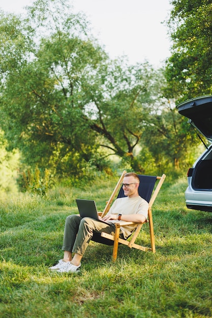 L'uomo libero professionista si siede su una sedia di legno nella natura e lavora online su un laptop Un uomo viaggia e lavora a distanza su un computer portatile Lavoro d'ufficio nella natura Vacanze