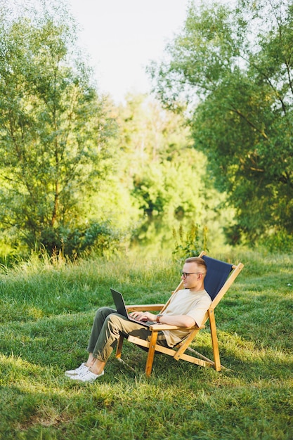 L'uomo libero professionista si siede su una sedia di legno nella natura e lavora online su un laptop Un uomo viaggia e lavora a distanza su un computer portatile Lavoro d'ufficio nella natura Vacanze