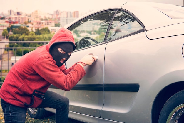 L'uomo mascherato si accovaccia e rompe l'auto di qualcuno guardando lo spettatore. Il ladro in felpa con cappuccio e faccia nascosta tenta di scassinare il veicolo parcheggiato fissando gli occhi alla telecamera. Il malfattore danneggia la serratura dell'auto.