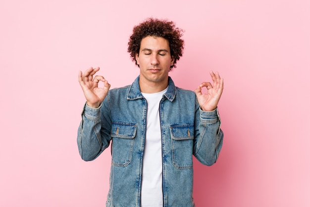 L'uomo maturo riccio che indossa una giacca di jeans contro la parete rosa si rilassa dopo una dura giornata di lavoro, sta eseguendo yoga.