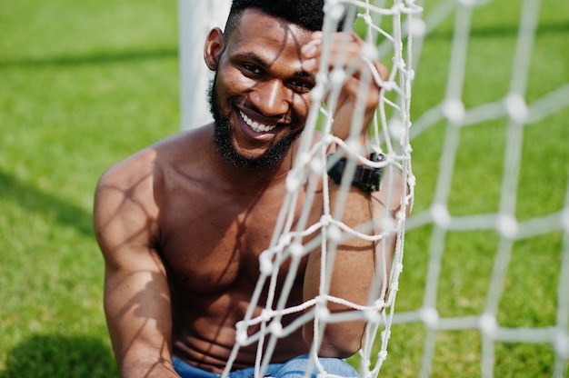 L'uomo muscoloso sexy bello del torso nudo alla tuta dei jeans ha posato alle porte di calcio del agasinst dell'erba verde del campo dello stadio