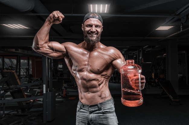 L'uomo muscoloso sta in palestra con un'enorme bottiglia di nutrizione sportiva. Concetto di fitness e bodybuilding. Tecnica mista