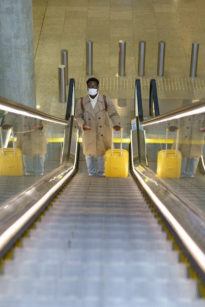 L'uomo nero con la valigia si trova sulla scala mobile in aeroporto indossa la maschera per il viso durante la pandemia covid-19.