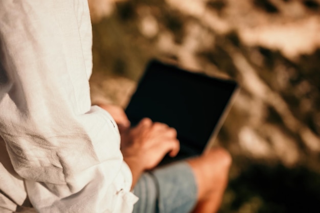 L'uomo nomade digitale con il cappello un uomo d'affari con un laptop si siede sulle rocce in riva al mare durante il tramonto