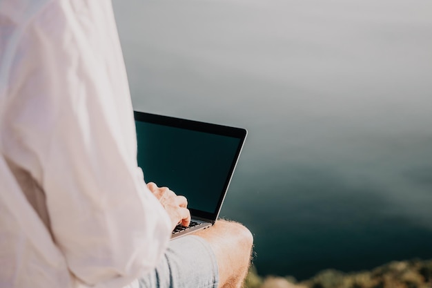 L'uomo nomade digitale con il cappello un uomo d'affari con un laptop si siede sulle rocce in riva al mare durante il tramonto