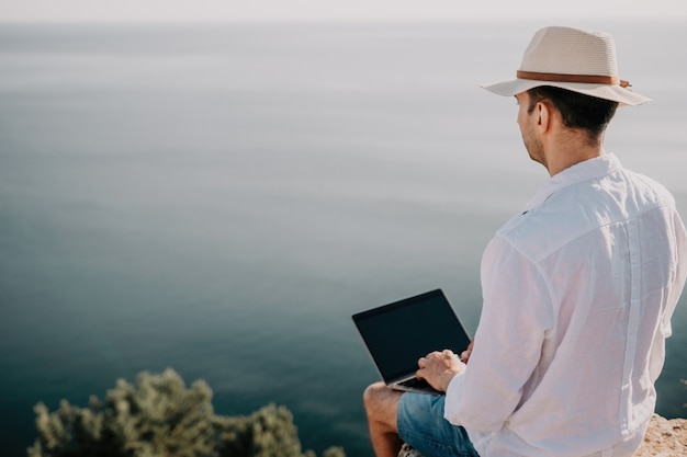 L'uomo nomade digitale con il cappello un uomo d'affari con un laptop si siede sulle rocce in riva al mare durante il tramonto