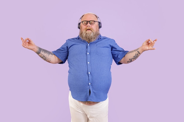 L'uomo obeso barbuto maturo tranquillo in camicia attillata medita mentre ascolta musica rilassante con le cuffie su sfondo viola in studio