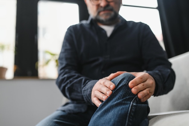 L'uomo più anziano è seduto sul divano di casa ha dolore all'articolazione del ginocchio che tiene il concetto di artrosi della gamba