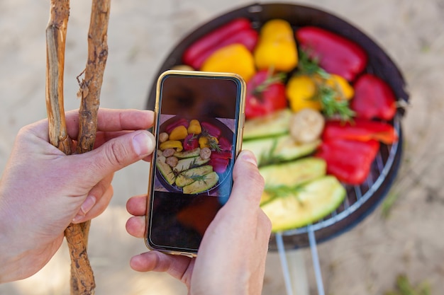 L'uomo prende le immagini sul telefono verdure grigliate. Verdure rosse e gialle peperoni, funghi e zucchine arrostite su un barbecue rotondo