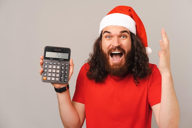L'uomo scioccato che indossa il cappello di Natale sta gesticolando mentre mostra la calcolatrice