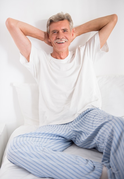 L'uomo senior allegro sta sedendosi a letto e sta esaminando la macchina fotografica.