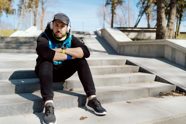 L'uomo serio si siede sui gradini nel parco con le braccia incrociate ascolta la musica con le cuffie wireless