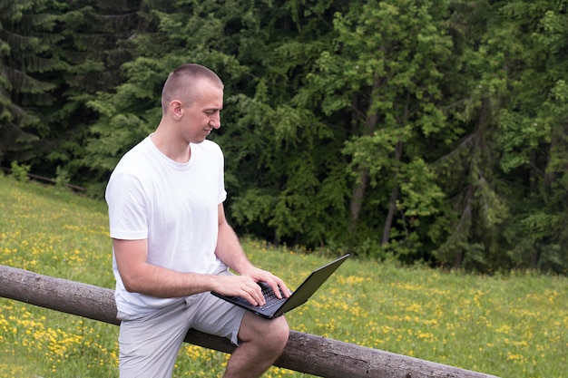 L'uomo si siede su una staccionata di legno e lavora con un computer portatile vicino al campo e alla foresta di conifere