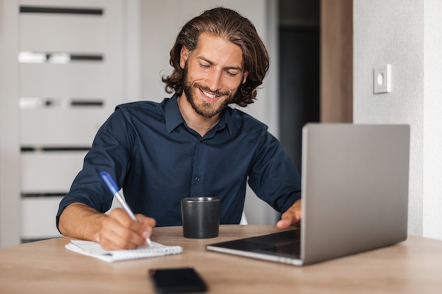 L'uomo soddisfatto con un sorriso lavora su un laptop e scrive su un notebook