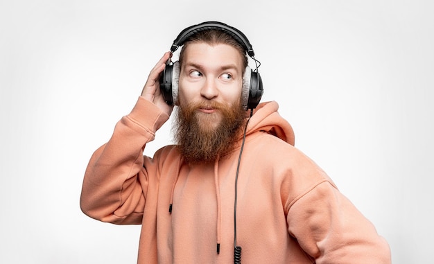 L'uomo sorpreso bello scandinavo guarda di lato e ascolta la musica in cuffie professionali isolate su sfondo grigio Ragazzo felice con la barba di capelli rossi Moderne tecnologie digitali