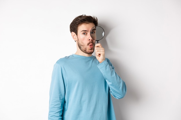 L'uomo sorpreso ha trovato qualcosa, guardando attraverso la lente d'ingrandimento con il viso stupito, in piedi su sfondo bianco.