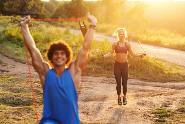 L'uomo sportivo caucasico sorridente con capelli ricci che fanno l'allungamento si esercita con la corda in natura nel giorno di estate soleggiato. In background donna saltando la corda. Messa a fuoco selettiva sulla donna.