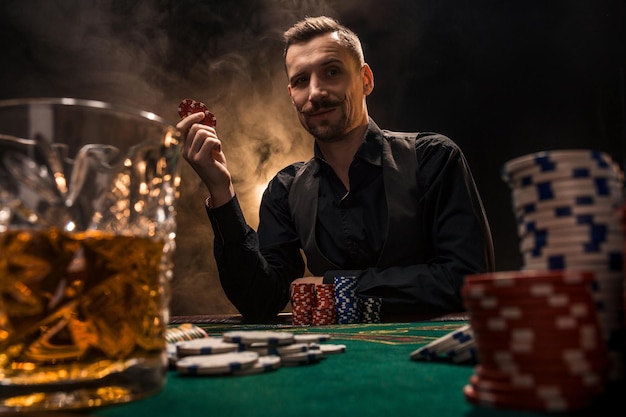 L'uomo sta giocando a poker con un sigaro e un whisky. Un uomo che vince tutte le fiches sul tavolo con un denso fumo di sigaretta. Il concetto di vittoria. Bicchiere con whisky e pila di patatine in primo piano