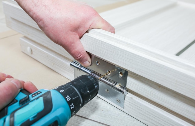 L'uomo sta installando le porte Il falegname sta facendo dei fori con il trapano nei cardini Lavori di riparazione Manutenzione nell'appartamento