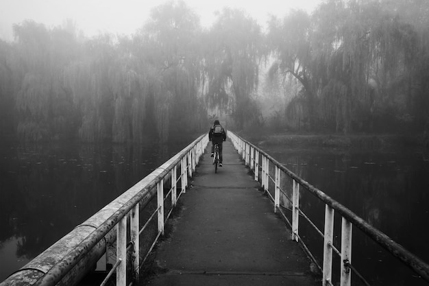 L'uomo sta pedalando attraverso il vecchio ponte arrugginito sul lago in una mattina presto nebbiosa