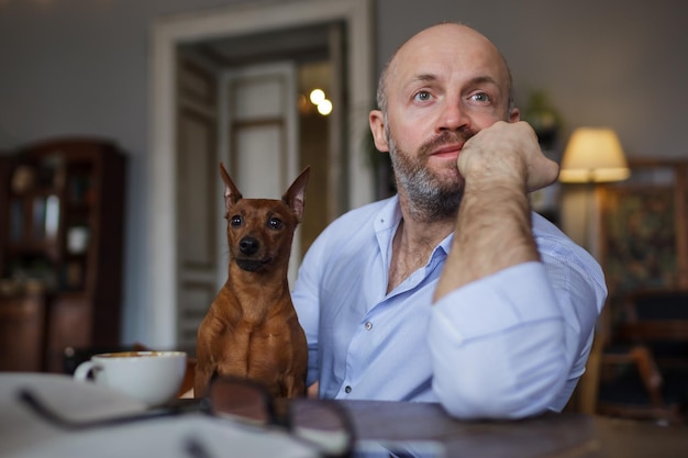 L'uomo sta riposando con il suo cane Un libro aperto e bicchieri giacciono sul tavolo Lui distoglie lo sguardo sognante