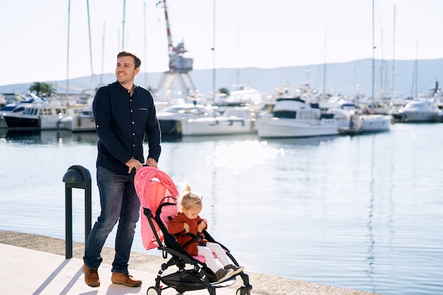 L'uomo sta spingendo un passeggino rosa con sua figlia di 2 anni dentro su un molo in una giornata di sole.