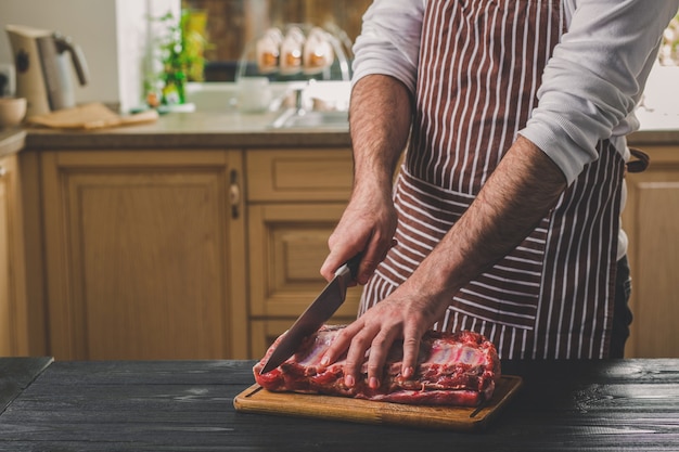 L'uomo taglia un pezzo di carne fresca su un tagliere di legno nella cucina di casa. Un uomo con un grembiule a righe con un grosso coltello in mano