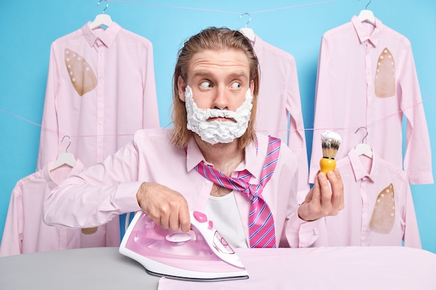 l'uomo tiene il pennello da barba applica la schiuma gel stira i vestiti si prepara per l'appuntamento vuole avere un aspetto brillante posa su camicie stirate sui ganci essere occupato dopo aver dormito