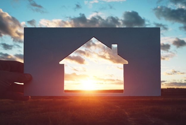 L'uomo tiene un pezzo di carta con una casa ritagliata contro il tramonto. Casa in natura, viaggi e alloggio. Concetto.