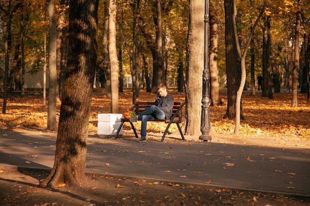 L'uomo triste è seduto su una panchina in un parco cittadino autunnale con un mazzo di fiori e aspetta a lungo con un'espressione insoddisfatta sul viso. Un appuntamento fallito